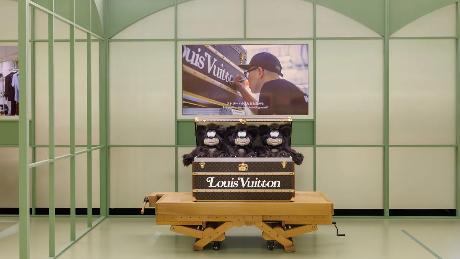 Louis Vuitton trunk with plush toys displayed open on a wooden stand. Background features a screen showing a person working on a trunk, creating a luxury and artistic ambiance.
