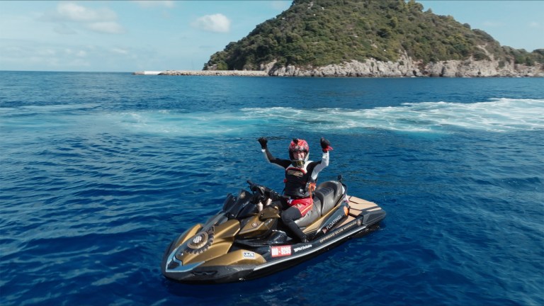 Person on a jet ski poses triumphantly on vibrant blue ocean, wearing a helmet and suit. Rocky, tree-covered island in background under a clear sky.