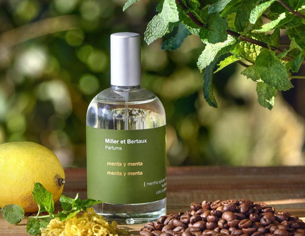 A bottle of Miller et Bertaux "menta y menta" perfume stands on a wooden surface with lemon, coffee beans, mint leaves, and lemon zest around it.