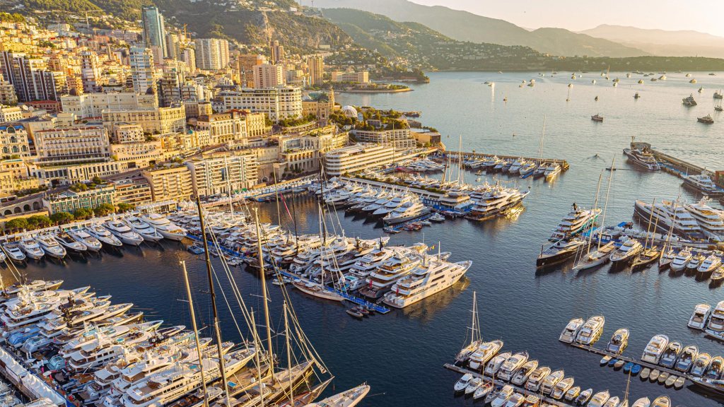 Aerial view of a bustling Monaco marina with numerous yachts in the foreground, set against a coastal cityscape at sunset. Hills loom in the background.