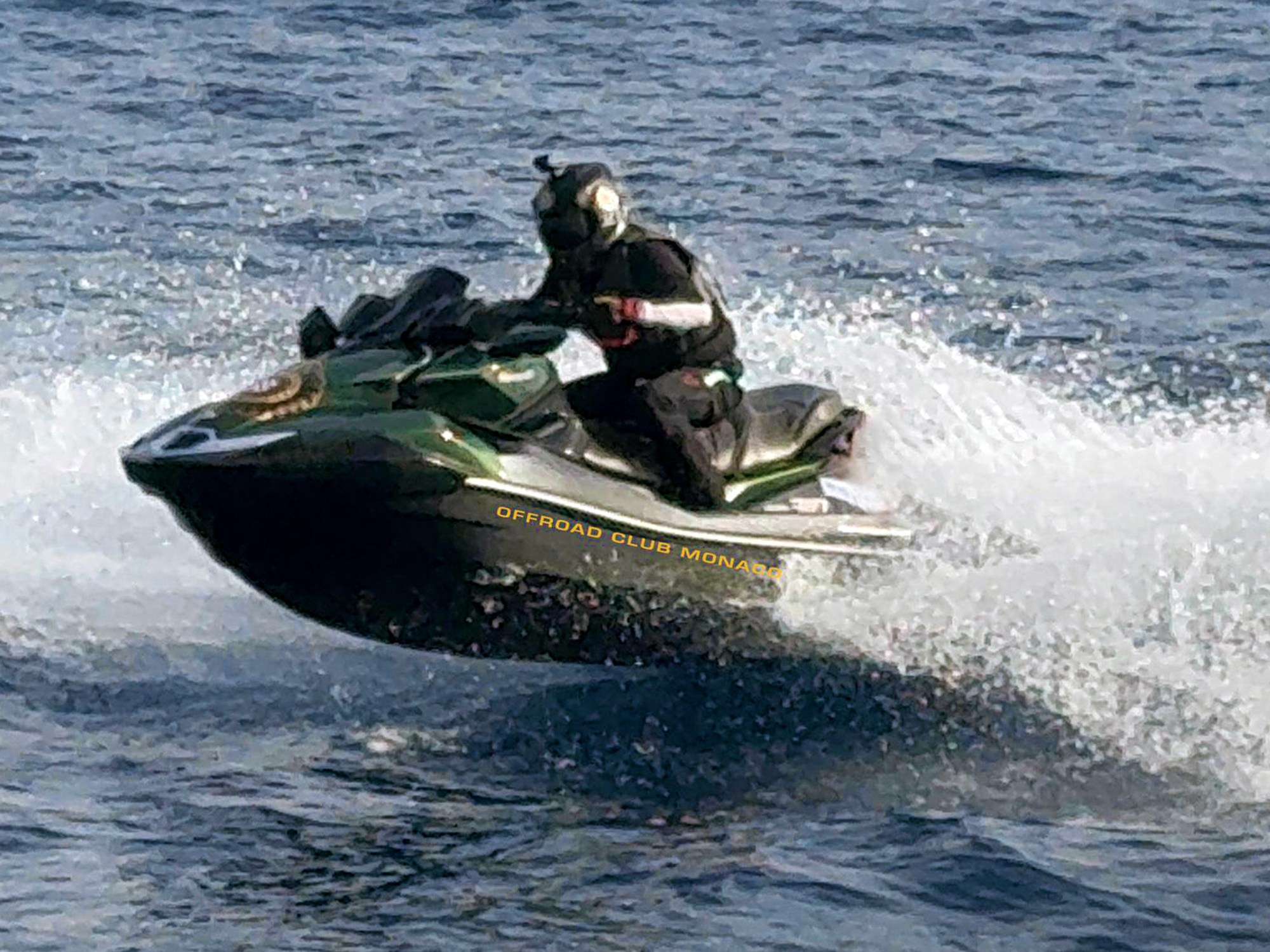 A person in black gear rides a green jet ski over the ocean, creating splashes. The scene conveys excitement and speed on a sunny day.