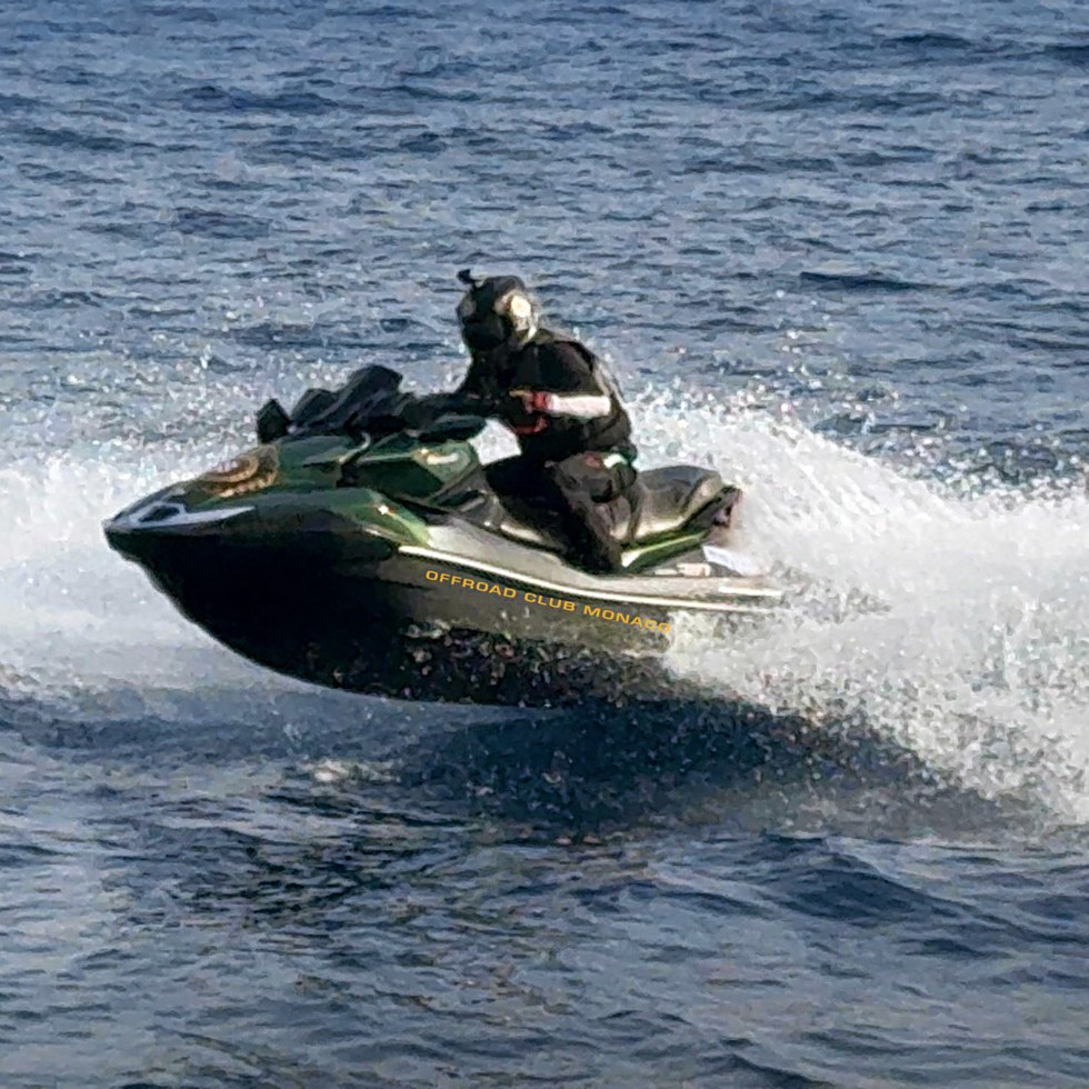 A person in black gear rides a green jet ski over the ocean, creating splashes. The scene conveys excitement and speed on a sunny day.