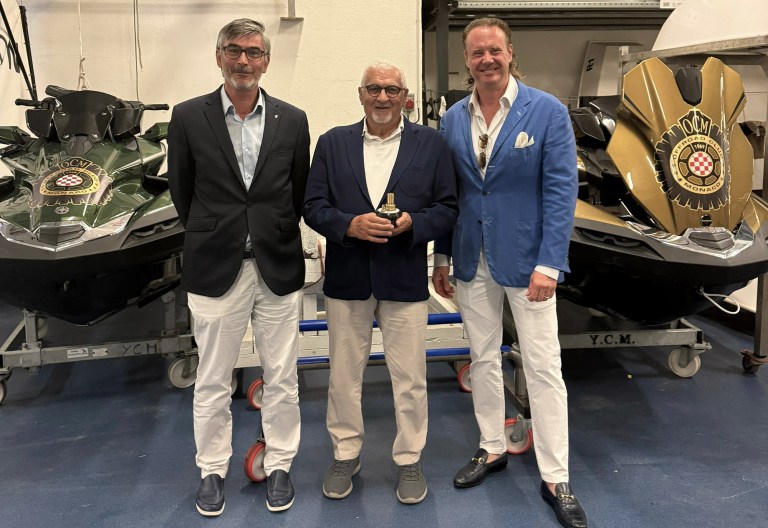 Three men stand together in a workshop between two luxury watercraft. They are smiling, dressed in smart casual blazers and white trousers. The mood is celebratory.