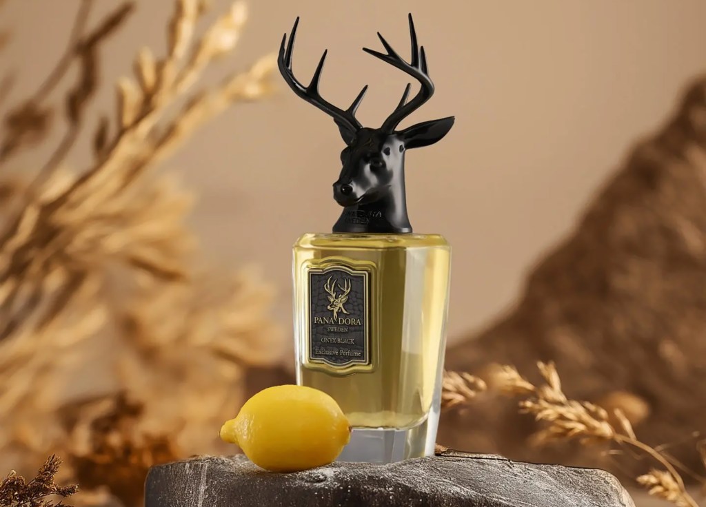 Perfume bottle with a black deer head cap on a stone surface, flanked by yellow lemons. Background features soft, earthy tones and dried plants.