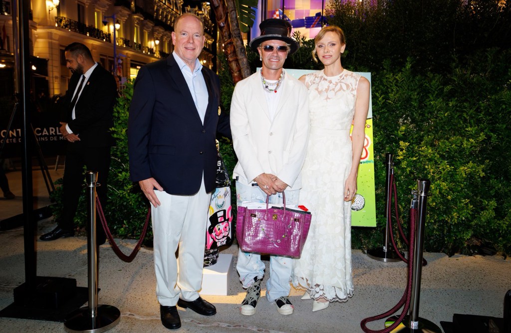 Prince Albert II, Alec Monopoly, and Princess Charlene pose together at the 2025 Monaco Cup event. Alec Monopoly wears a stylish white outfit and carries a vibrant handbag, while Princess Charlene dazzles in an elegant white lace dress.
