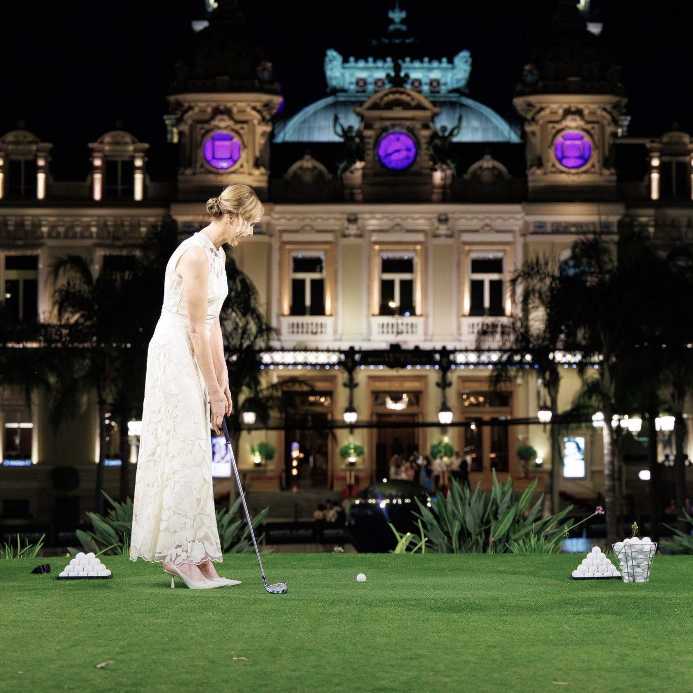 Princess Charlene in an elegant Elie Saab white dress plays mini-golf on a lush green lawn. Behind her is an illuminated, ornate Casino de Monte-Carlo with purple-lit clocks, creating a sophisticated nighttime ambiance.