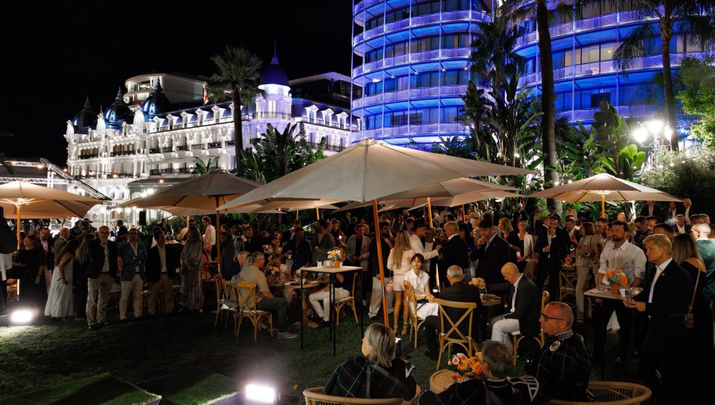 A crowded outdoor venue at night during the 2025 Monaco Cup, featuring elegantly dressed guests mingling under large umbrellas with a beautifully lit historic building in the background.