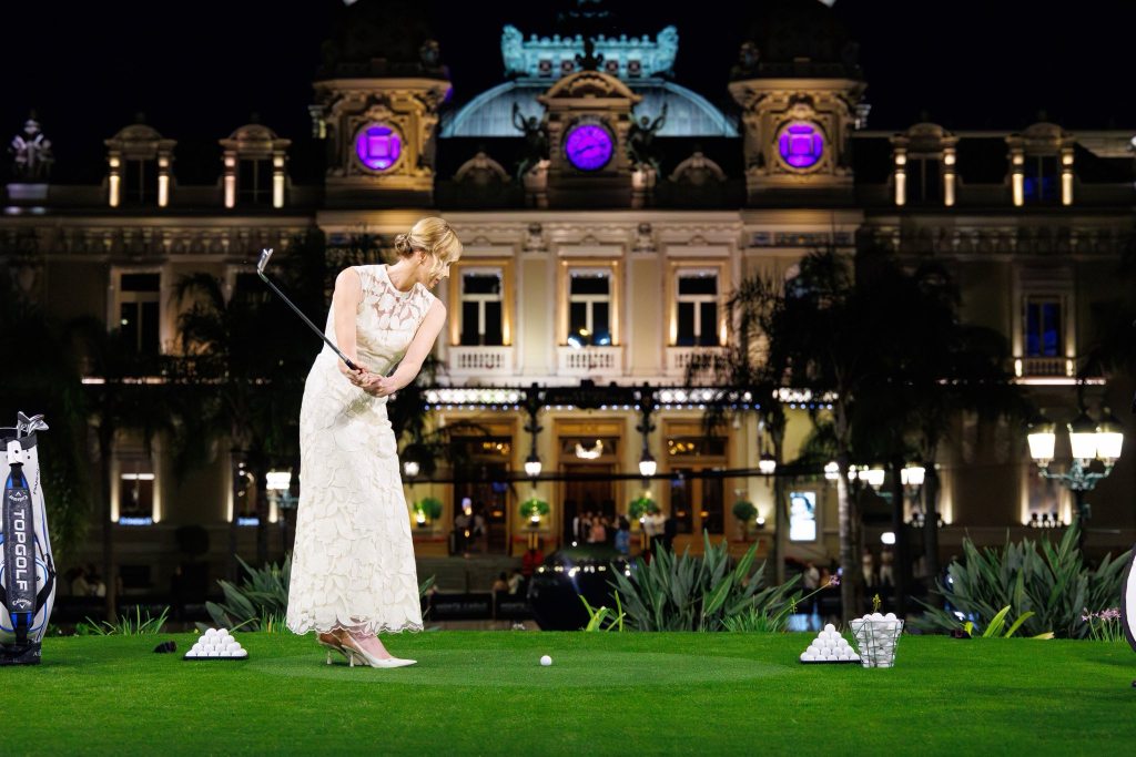 A woman wearing an elegant white lace dress is preparing to swing a golf club at the Monaco Cup event, with the illuminated backdrop of a grand building in the evening.