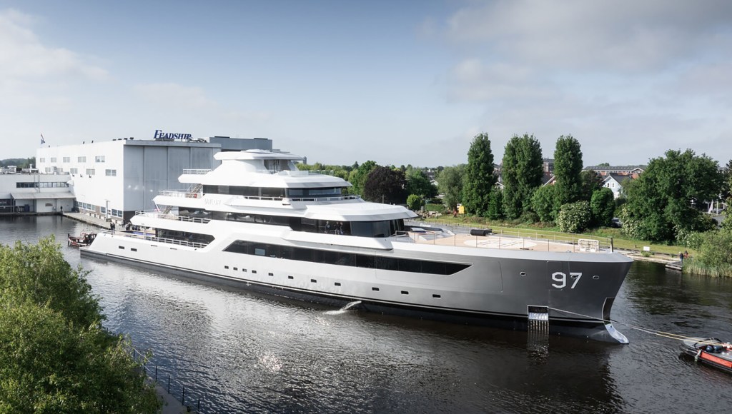 Luxurious yacht with sleek silver design and multiple decks is docked near a modern shipyard building. Trees and blue sky in the background add serenity.