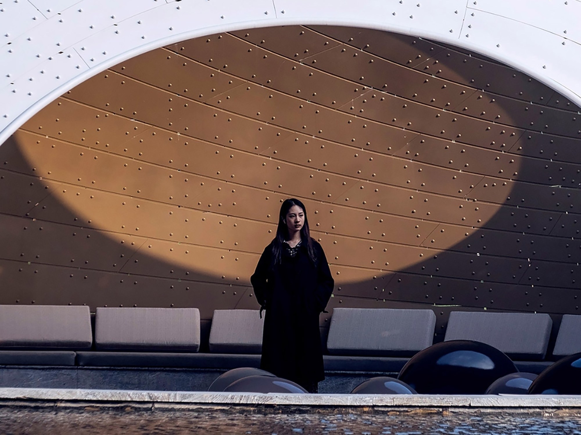 A woman stands in a dramatic setting, framed by an architectural arch with a dotted pattern. She's near a reflective pool; the mood is serene and contemplative.