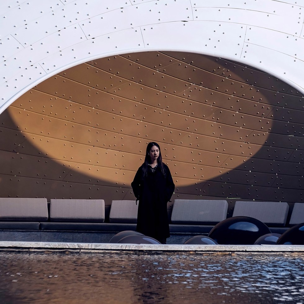 A woman stands in a dramatic setting, framed by an architectural arch with a dotted pattern. She's near a reflective pool; the mood is serene and contemplative.
