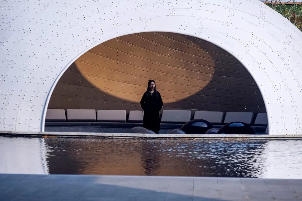 A person stands under a large, modern white arch by a reflective pool, showing a serene yet contemplative scene. Soft seating lines the background.