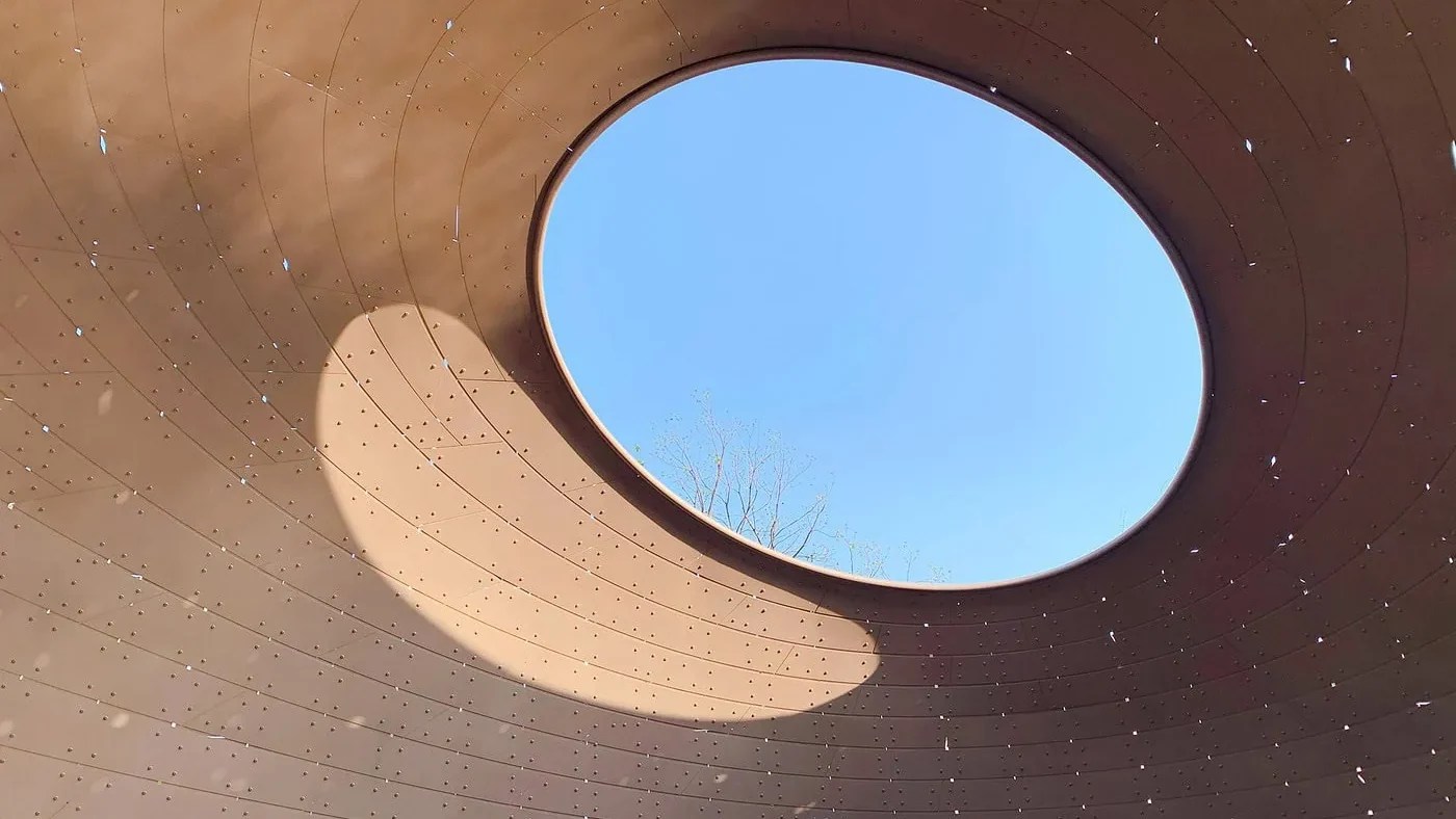 Circular skylight in a textured dome, framing a bright blue sky. Light and shadows create a peaceful, open ambiance within the space.