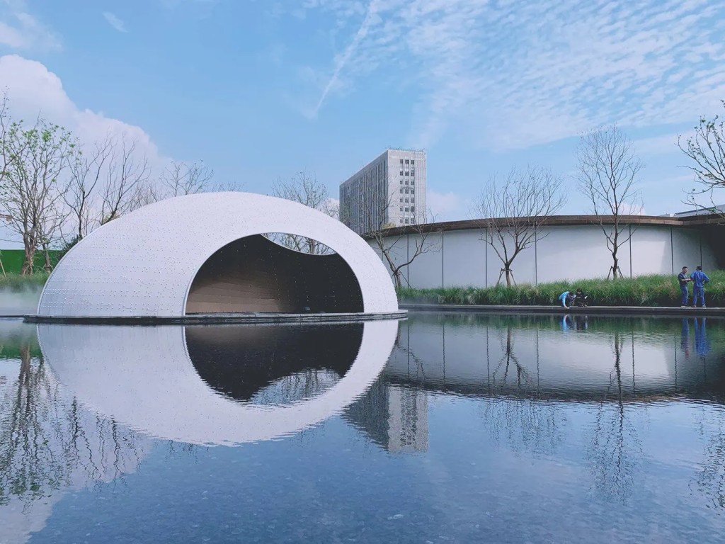 A white domed structure with a large entrance reflects in a calm pond under a clear blue sky. Nearby, people stand by a curved building and trees.