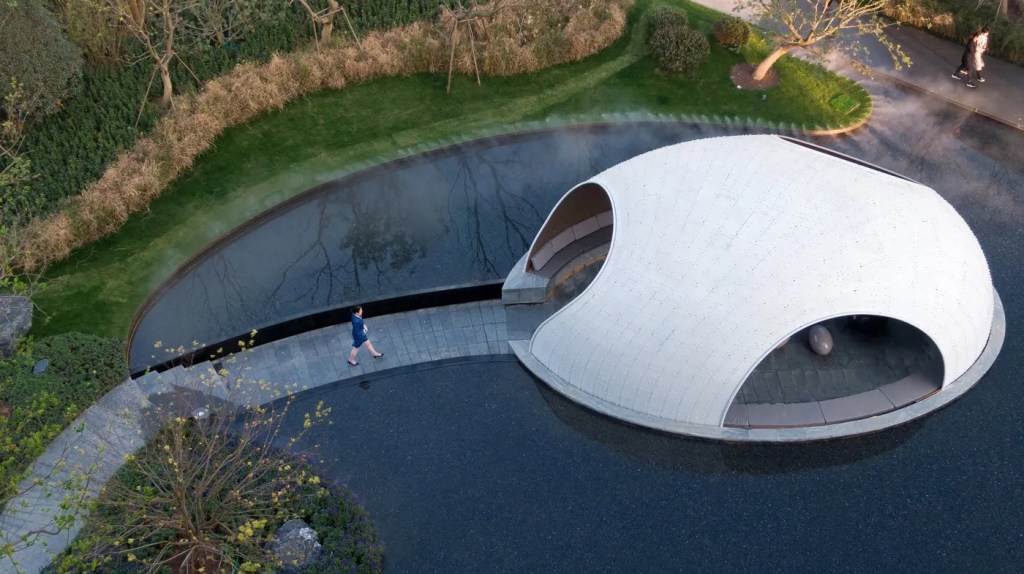 A person walks on a curved path beside a large, dome-shaped structure surrounded by a shallow pond. The setting is tranquil and modern.