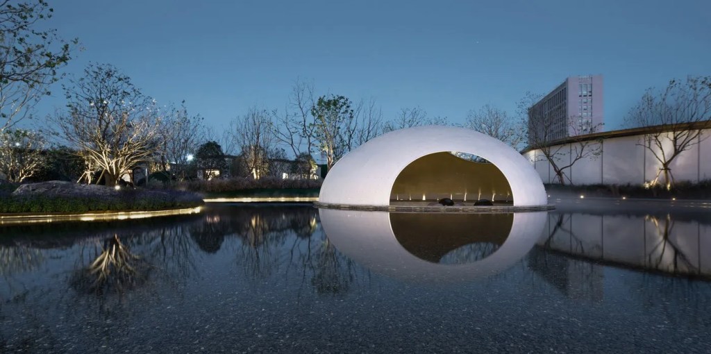A serene nighttime scene shows a white dome structure with an archway, reflected in a calm pond. Sparse trees and a building are in the background.