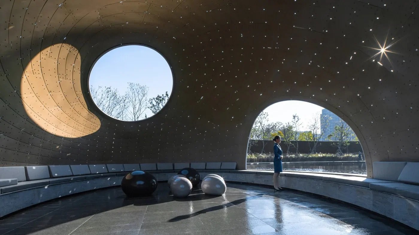 Futuristic room with large circular windows and star-shaped lighting, featuring a person observing three shiny, spherical sculptures on a glossy floor.
