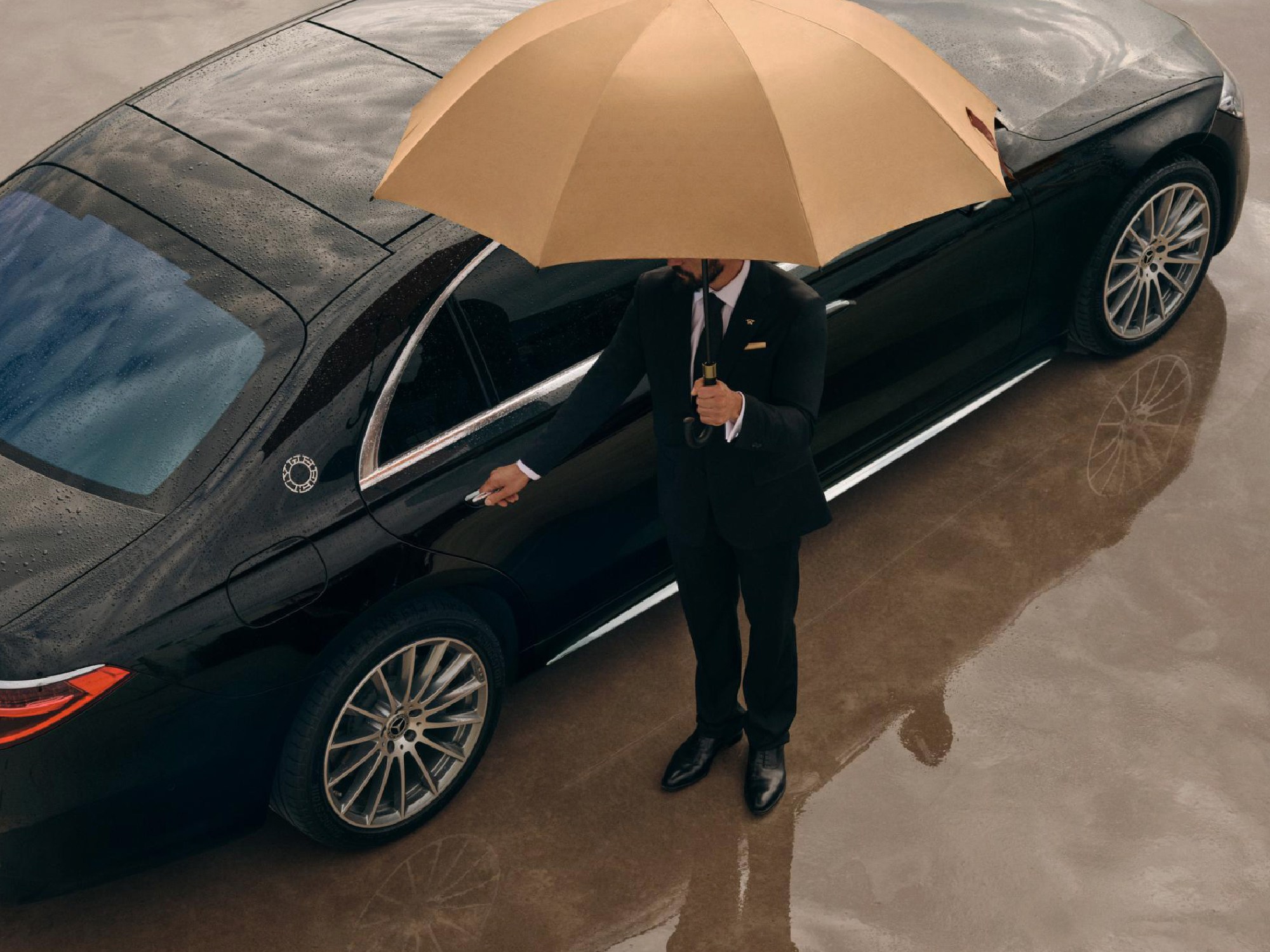 wheely personal chauffer opens door of black maybach and holds beige umbrella to protect from the rain