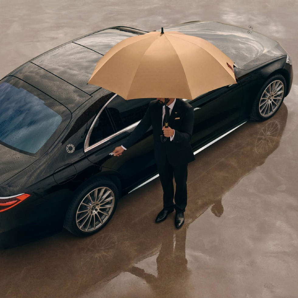 wheely personal chauffer opens door of black maybach and holds beige umbrella to protect from the rain